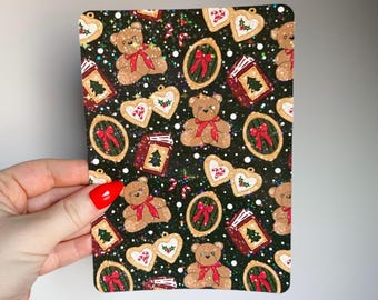 May include: A rectangular card with a repeating pattern of teddy bears with red bows, heart-shaped ornaments, framed red bows, and books with Christmas trees on a dark green background. The card is held by a hand with a red painted nail.
