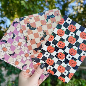 May include: Three Halloween themed decorative sheets with a glittery finish. One sheet features a pink and white checkered pattern with white flowers and bats. Another sheet features a brown and white checkered pattern with bats and stars. The third sheet features a black and white checkered pattern with orange pumpkins and stars.