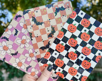 Pode incluir: Três folhas decorativas com tema de Halloween com acabamento brilhante. Uma folha apresenta um padrão xadrez rosa e branco com flores brancas e morcegos. Outra folha apresenta um padrão xadrez marrom e branco com morcegos e estrelas. A terceira folha apresenta um padrão xadrez preto e branco com abóboras laranja e estrelas.