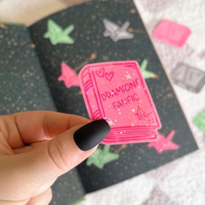 May include: Pink book-shaped sticker with glitter and the text "DRAMIIONE FANFIC" and two hearts.