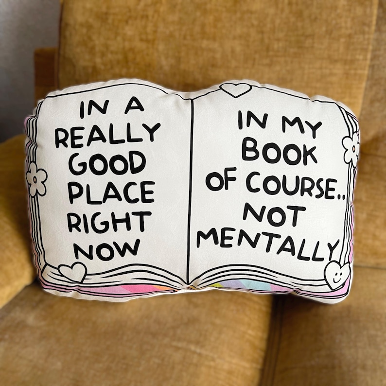 May include: A white book-shaped pillow with black text that reads "In a really good place right now" on the left side and "In my book of course... not mentally" on the right side. The pillow has pink and rainbow accents.