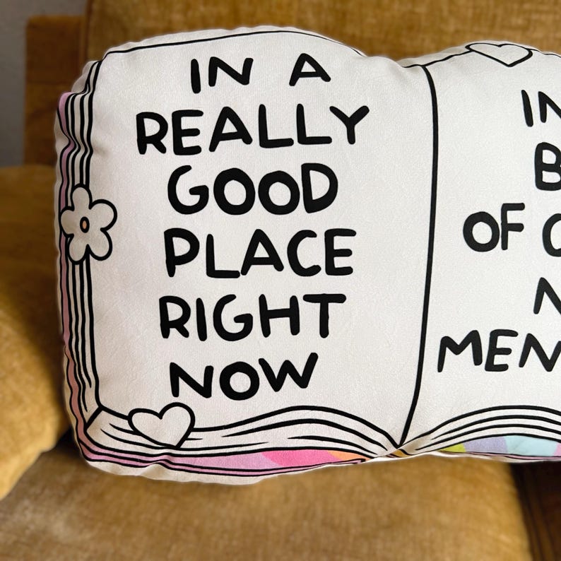 May include: A white book-shaped pillow with black text that reads "In a really good place right now". The pillow has a pink and rainbow design on the bottom edge.