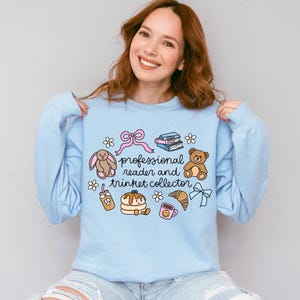 May include: A light blue crewneck sweatshirt featuring a central graphic with the text "professional reader and trinket collector" surrounded by illustrations of a pink bow, books, a brown bunny, a teddy bear, pancakes, a croissant, and a mug.