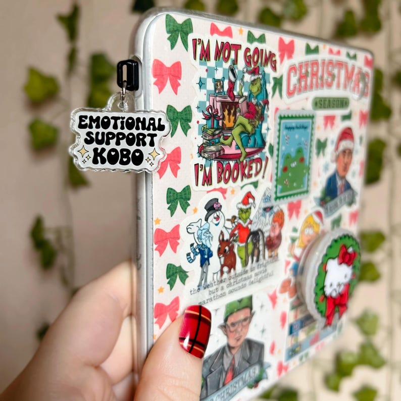 May include: A white phone case with a variety of colorful stickers, including a Grinch character, a snowman, and a Christmas-themed design. The case also has a clear plastic charm with the text "Emotional Support Kobo" in black lettering.