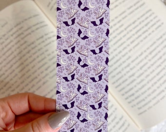 Puede incluir: Un marcapáginas morado y blanco con un patrón repetido de murciélagos, lunas y búhos leyendo libros.