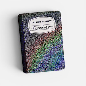 Könnte beinhalten: Eine rechteckige Kindle-Hülle mit einer leuchtenden, mehrfarbigen holografischen Glitzer-Oberfläche. Mittig oben befindet sich ein weißes Etikett mit schwarzem Rand, das den Text "THIS KINDLE BELONGS TO" und den handgeschriebenen Namen "Amber" enthält.