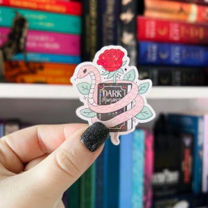 Könnte beinhalten: Eine rosa Schlange windet sich um ein schwarzes Buch mit dem Schriftzug "DARK ROMANCE" auf dem Einband. Hinter dem Buch befindet sich eine rosa Rose mit grünen Blättern. Der Sticker ist auf weißem Hintergrund.