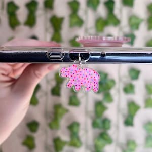 Puede incluir: Un charm de teléfono rosa con forma de oso, cubierto de lunares de colores, cuelga de un teléfono. El charm está sujeto al teléfono con un lazo de metal plateado. También se ve un soporte de teléfono rosa.