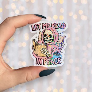 May include: A holographic sticker showing a skeleton in a pink hoodie reading a book, with the text "LET ME READ IN PEACE". The skeleton is holding a book and a cup of coffee. The sticker has a glittery effect.