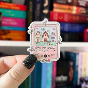 May include: A pink and white sticker with a glittery finish featuring a cartoon illustration of three houses with a heart above each. The sticker has a music player design with the text "Small Town Romance Audiobook Series" and a play button.