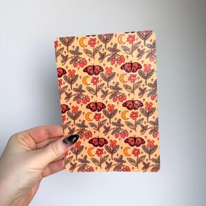 May include: A rectangular notebook with a repeating pattern of butterflies, flowers, moons, and berries in shades of orange, red, and brown on a light orange background. The notebook is held by a hand.