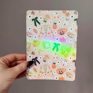 May include: A rectangular, patterned item featuring a light pink background with a repeating pattern of pumpkins, sweaters, mugs, bows, and flowers. The design has a fall or autumn theme, with a touch of Halloween. The item is held by a hand.