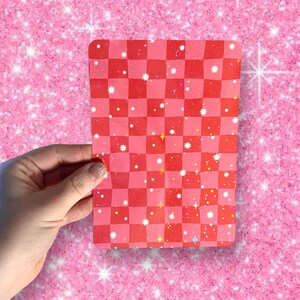 Puede incluir: Una pegatina a cuadros rojos y blancos con un acabado brillante. La pegatina tiene un fondo rosa con estrellas blancas y purpurina de colores.