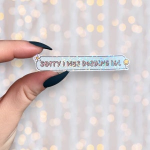 May include: A holographic sticker with a blue chat bubble and the text "Sorry I was reading lol" in pink. A small white daisy with a yellow center is in the upper left corner of the chat bubble. A yellow smiley face is in the upper right corner of the chat bubble.