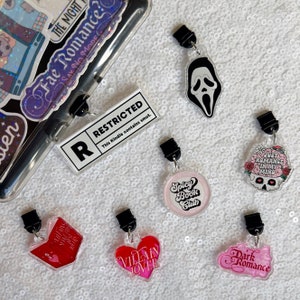 Op de afbeelding: Een set van tien kleurrijke boekencharms met verschillende ontwerpen, waaronder een schedel met bloemen, een hart met de tekst "Villain Lover", een boek met de tekst "Villains Make the Best", een spookgezicht, een cirkel met de tekst "Spicy Book Club" en een cirkel met de tekst "Dark Romance". De charms zijn bevestigd aan doorzichtige plastic clips.