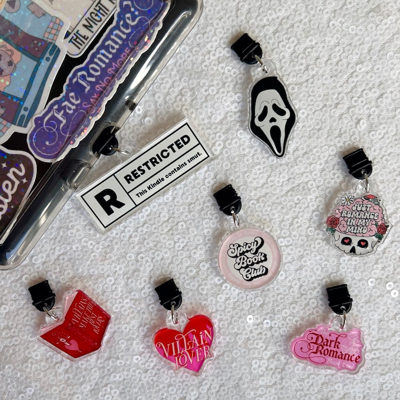 Op de afbeelding: Een set van tien kleurrijke boekencharms met verschillende ontwerpen, waaronder een schedel met bloemen, een hart met de tekst "Villain Lover", een boek met de tekst "Villains Make the Best", een spookgezicht, een cirkel met de tekst "Spicy Book Club" en een cirkel met de tekst "Dark Romance". De charms zijn bevestigd aan doorzichtige plastic clips.