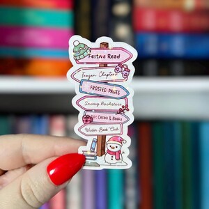 May include: A whimsical sticker featuring a signpost design with winter-themed destinations. The signpost has arrows pointing to "Festive Read," "Frozen Chapters," "Frosted Pages," "Snowy Bookstore," "Hot Cocoa & Books," and "Winter Book Club." A snowman and books are at the base.