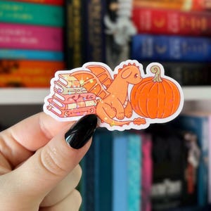 May include: A sticker featuring an orange dragon sitting next to a pumpkin and a stack of books. The books are pink and orange with gold accents. The dragon has orange wings and a cute expression. The sticker is held by a hand with a black nail.