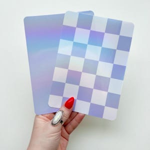May include: Two rectangular, iridescent cards. One card has a checkerboard pattern in shades of lavender and white. The other card has a solid lavender color with a subtle rainbow sheen. The cards are held by a hand.