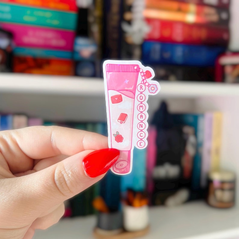 Puede incluir: Una pegatina rosa con forma de tubo con la palabra "ROMANCE" escrita verticalmente. La pegatina tiene peque&ntilde;as ilustraciones de un coraz&oacute;n, un libro y una fresa. El fondo est&aacute; borroso, mostrando una estanter&iacute;a con libros.
