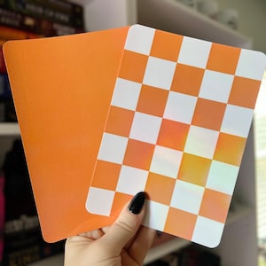 Könnte beinhalten: Zwei Karten, eine einfarbig orange und die andere mit einem karierten Muster aus orange und weißen Quadraten. Die Karten werden vor einem verschwommenen Hintergrund gehalten. Die Karten sind rechteckig mit abgerundeten Ecken.