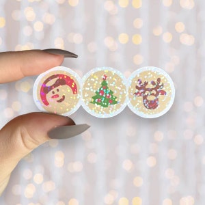 May include: A horizontal strip of three circular stickers featuring a tan background with a holographic glitter finish. The left sticker shows a red and white Santa hat, the center a green Christmas tree, and the right a brown reindeer face.