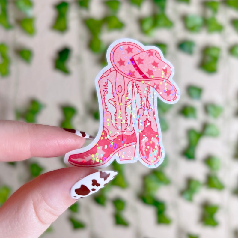 May include: Pink glitter cowboy boots with a pink glitter cowboy hat on top. The boots and hat are on a white background.