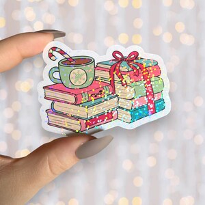 May include: A holographic sticker featuring a stack of books tied with a red ribbon, a mug of hot chocolate with a candy cane, and a snowflake design. The books have pink, green, and blue covers. The sticker has a glittery, iridescent effect.
