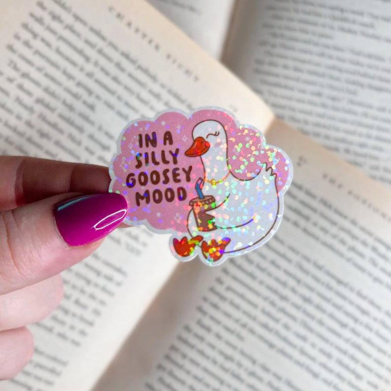 May include: A holographic sticker with a pink background and a white goose holding a cup of coffee. The sticker says "In a silly goosey mood".