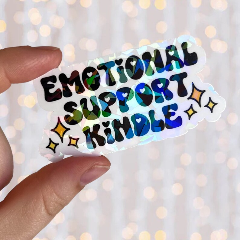 May include: A holographic sticker with the text "Emotional Support Kindle" in black lettering. The sticker has a rainbow iridescent background with small hearts and stars.