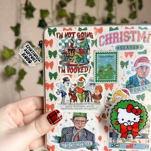 May include: A close-up of a clear plastic binder with a variety of colorful Christmas-themed stickers. The stickers feature characters like the Grinch, the Abominable Snowman, and Hello Kitty. The text on the stickers includes "I'm not going", "I'm booked", "Christmas Season", "Happy Holidays", and "It is Christmas".