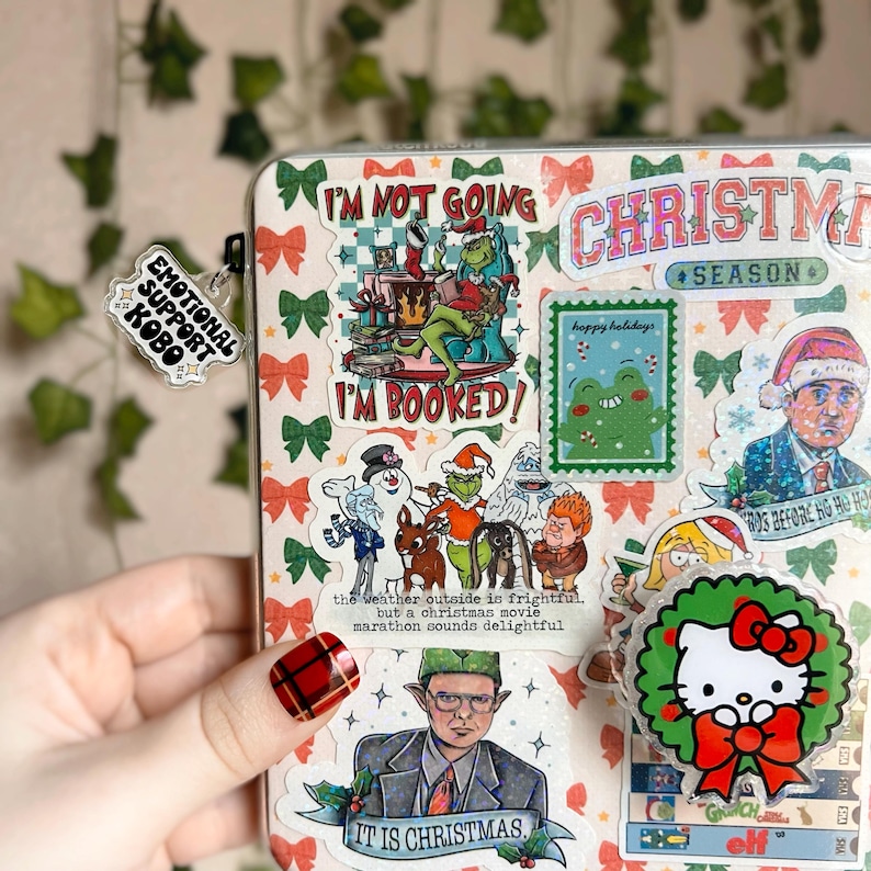 May include: A close-up of a clear plastic binder with a variety of colorful Christmas-themed stickers. The stickers feature characters like the Grinch, the Abominable Snowman, and Hello Kitty. The text on the stickers includes "I'm not going", "I'm booked", "Christmas Season", "Happy Holidays", and "It is Christmas".