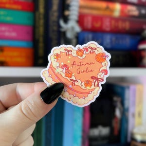 Puede incluir: Una pegatina en forma de coraz&oacute;n con un dise&ntilde;o de pastel naranja, con calabazas, setas y el texto "Autumn Girlie". La pegatina tiene un fondo naranja claro con un borde decorativo y lazos. La pegatina se sostiene en una mano.