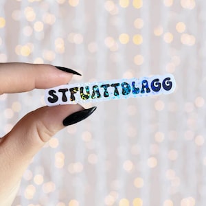 May include: A white sticker with the text "STFUATTDBLAGG" in black, glittery letters. The sticker is held in a hand with black nail polish.