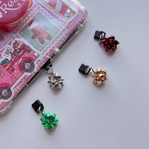 May include: A pink phone case with a Christmas theme, featuring a gingerbread man and a red truck. Four bow-shaped charms in silver, red, gold, and green are attached to black connectors, suggesting a festive accessory.