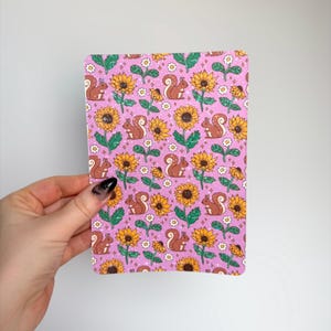 May include: A rectangular card with a repeating pattern of sunflowers and squirrels on a pink background. The sunflowers are yellow and brown, and the squirrels are brown. White daisy-like flowers are also scattered throughout the design.