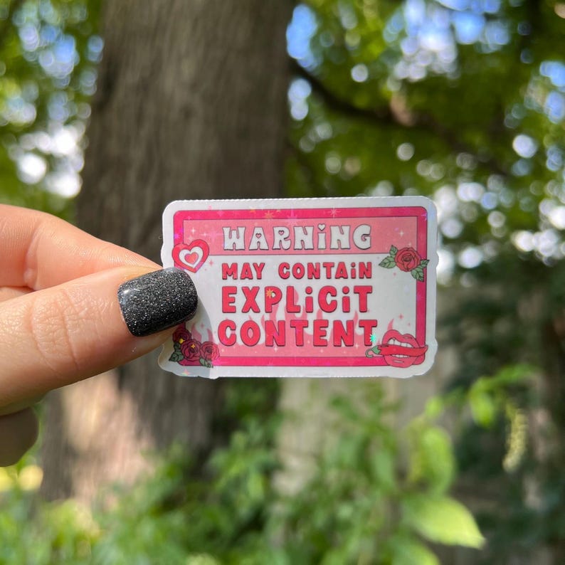 Op de afbeelding: Roze en witte waarschuwingsticker met de tekst "WARNING MAY CONTAIN EXPLICIT CONTENT" in roze en witte lettertypen. De sticker heeft een rood hart, rode rozen en een rood lippenontwerp.