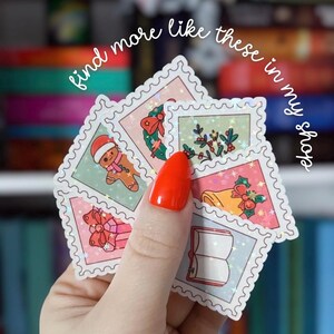 Puede incluir: Una mano sosteniendo una colecci&oacute;n de pegatinas con tem&aacute;tica navide&ntilde;a. Las pegatinas presentan dise&ntilde;os festivos como un hombre de jengibre, una campana y un regalo envuelto. El texto "find more like these in my shop" est&aacute; escrito en cursiva.
