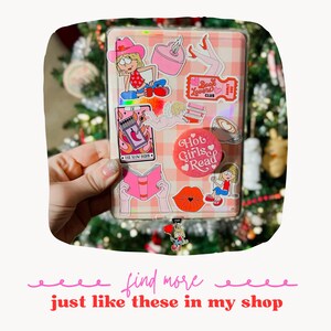 Puede incluir: Una libreta de notas a cuadros rosa y blanca con varias pegatinas, incluyendo una chica con un sombrero de vaquero, un libro, un coraz&oacute;n y una taza de caf&eacute;. La libreta tiene un charm rosa adjunto. El texto "Book Lovers Club" y "Hot Girls Read" es visible en las pegatinas.