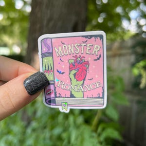 May include: A sticker of a book cover with a pink background and the text "Monster Romance". A green hand with a red heart is holding the heart in the center of the sticker.