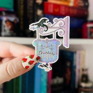 May include: A holographic sticker featuring a sign with the text "The Fantasy Reader" and a dragon design. The sign is in a blue and purple color scheme with a white border. The sticker is on a hand with a strawberry nail design.