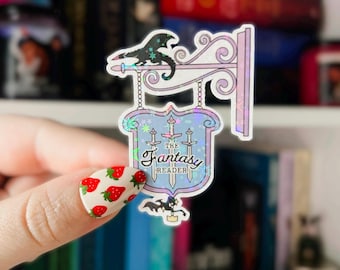 Può includere: Un adesivo olografico con un cartello con il testo "The Fantasy Reader" e un design di drago. Il cartello è in uno schema di colori blu e viola con un bordo bianco. L'adesivo è su una mano con un design di unghie a fragola.