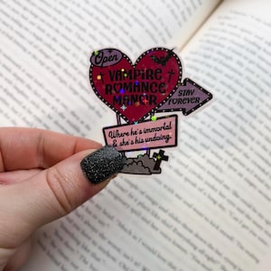 May include: A holographic sticker with a red heart-shaped sign that says "Vampire Romance Manor" and a purple arrow that says "Stay Forever". The sticker also has a smaller sign that says "Where he's immortal & she's his undoing."