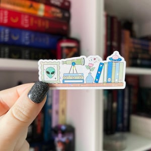 May include: A colorful sticker featuring a shelf with an alien portrait, telescope, books, a vase with flowers, and a UFO. The sticker has a holographic effect and is held by a hand with black glitter nail polish. The theme is space and reading.