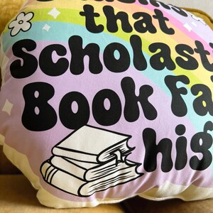 May include: A purple and white pillow with a rainbow and the text "Guess that Scholastic Book Fair is high" in black. A stack of books is illustrated in black and white.