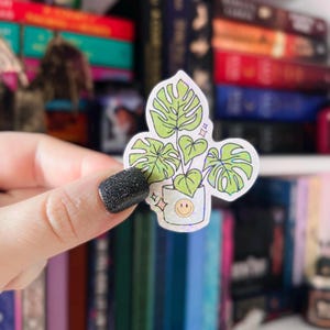 Pu&ograve; includere: Un adesivo olografico con una pianta di monstera verde in un vaso bianco con un viso sorridente. L'adesivo ha una finitura scintillante e iridescente.