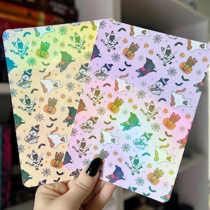 May include: Two Halloween-themed notebooks with colorful cat illustrations. The notebooks feature a repeating pattern of cats in witch hats, ghosts, pumpkins, spiderwebs, and bats. The background colors are a gradient of yellow, orange, and purple.