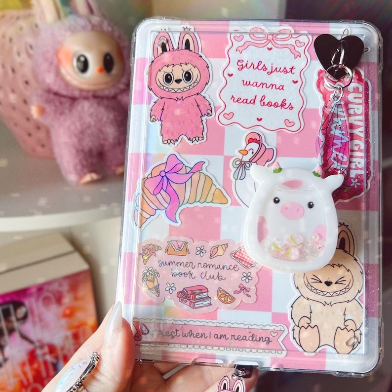 Puede incluir: Funda transparente para tel&eacute;fono con fondo a cuadros rosa y blanco, decorada con pegatinas de un conejito rosa, un cruas&aacute;n, una vaca y texto que dice "Girls just wanna read books" y "summer romance book club". Un charm de coraz&oacute;n negro y un llavero "CURVY GIRL" est&aacute;n adjuntos.