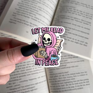 May include: A pink and white sticker with a skeleton wearing a pink hoodie and holding a book. The sticker says "Let me read in peace".