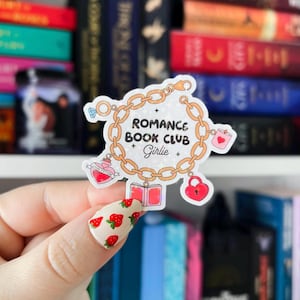 May include: A gold chain charm with a pink heart, a book, a bottle of potion, and a lock. The charm is surrounded by a white circle with the text "Romance Book Club Girlie".